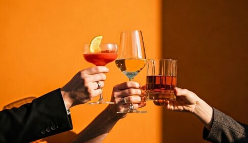 オレンジの壁の前でワインやウイスキーで乾杯する三人の手